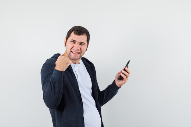 Jovem macho segurando o celular em t-shirt, jaqueta e parecendo com sorte. vista frontal.