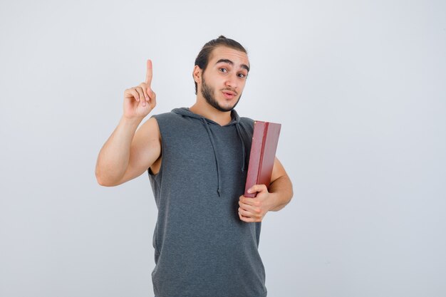 Jovem macho segurando o livro enquanto aparecendo apontando para cima com um capuz sem mangas e olhando confiante, vista frontal.