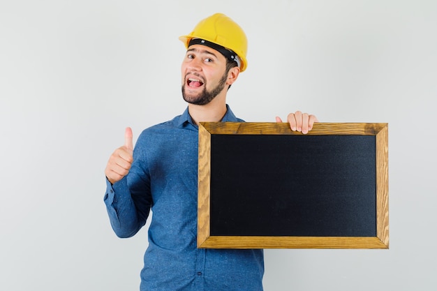 Jovem macho segurando o quadro-negro, aparecendo o polegar na camisa, capacete e olhando feliz. vista frontal.