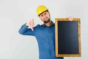 Foto grátis jovem macho segurando o quadro-negro, mostrando o polegar para baixo na camisa, capacete e parecendo descontente, vista frontal.