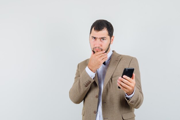 Jovem macho segurando o telefone móvel de terno e olhando pensativo, vista frontal.