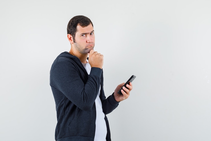 Foto grátis jovem macho segurando o telefone móvel em t-shirt, jaqueta e olhando pensativo, vista frontal.
