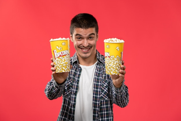 Jovem macho segurando pacotes de pipoca e sorrindo em um filme de cinema com parede vermelha clara