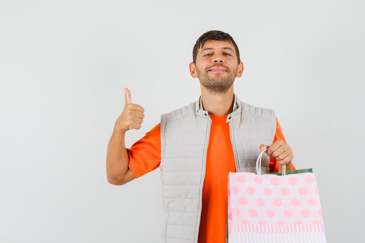 Foto grátis jovem macho segurando sacolas de compras, aparecendo o polegar em t-shirt, jaqueta e olhando jovial, vista frontal.