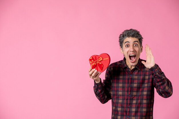 Jovem macho segurando um coração vermelho em forma de presente na parede rosa claro de frente