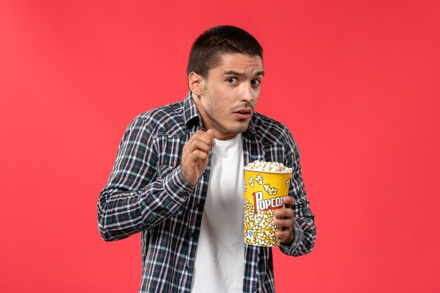 Jovem macho segurando um pacote de pipoca com expressão de medo na parede vermelha filme de cinema.