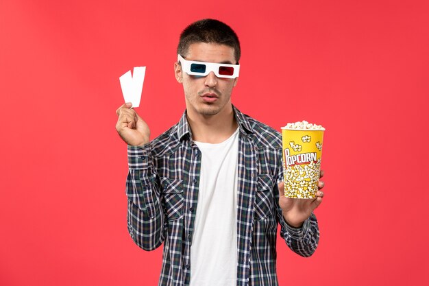 Jovem macho segurando um pacote de pipoca com ingressos para o filme de cinema de parede vermelho claro