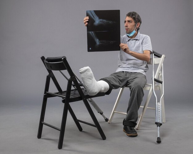 Jovem macho sentado com o pé quebrado segurando um raio-x na parede cinza.