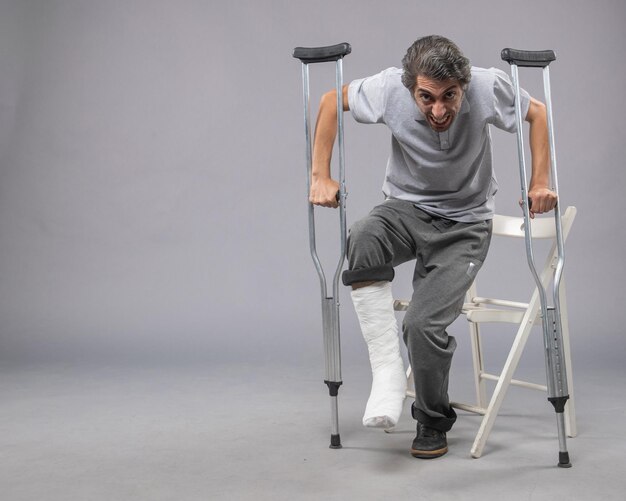 Jovem macho tentando se levantar com o pé quebrado usando muletas na parede cinza Pernas torção dor acidente pé quebrado