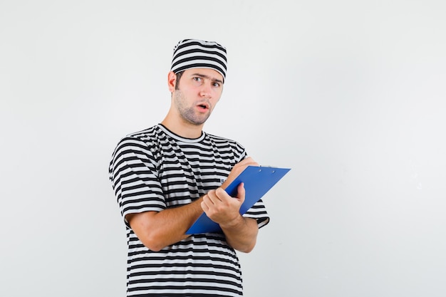 Jovem macho tomando notas na área de transferência em t-shirt, chapéu e olhando hesitante, vista frontal.