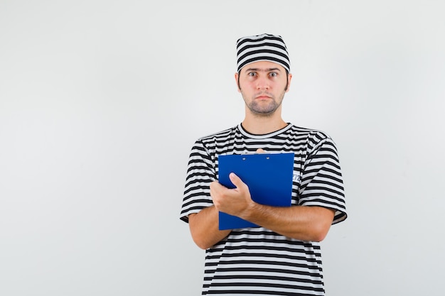 Jovem macho tomando notas na área de transferência em t-shirt, chapéu e parecendo perplexo. vista frontal.