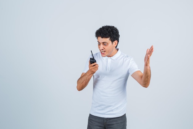 Jovem macho usando o telefone walkie-talkie da polícia em camiseta branca, calça e parecendo animado. vista frontal.