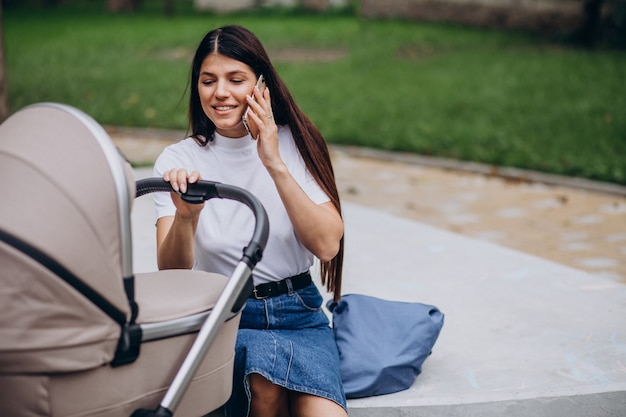 Foto grátis jovem mãe caminhando no parque com um carrinho de bebê e falando ao telefone