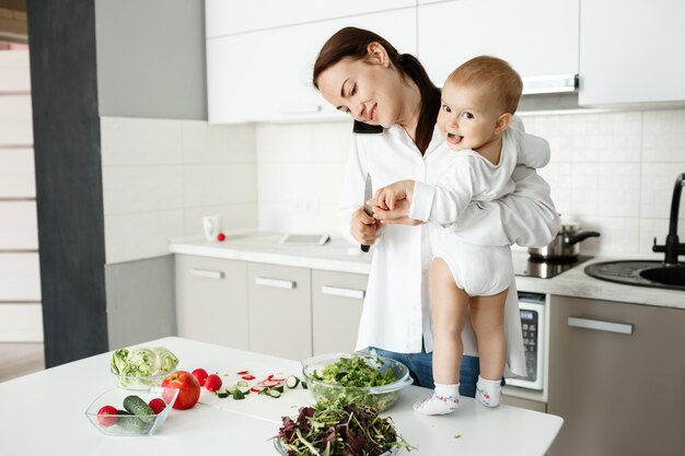 Jovem mãe cuidando de uma criança, falando ao telefone e cozinhando ao mesmo tempo