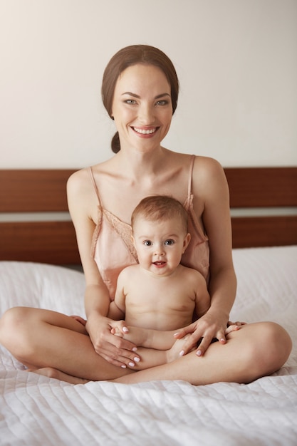 Jovem mãe feliz linda em roupa de noite e seu bebê recém-nascido sentado na cama de manhã sorrindo jogando juntos.