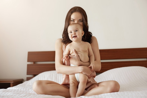 Jovem mãe feliz linda em roupa de noite e seu bebê recém-nascido sentado na cama de manhã sorrindo jogando juntos