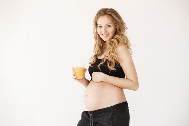 jovem mãe loira europeia com cabelos longos encaracolados em roupa confortável, sorrindo, bebendo suco de laranja pela manhã, mostrando sua barriga grande no nono mês de gravidez.
