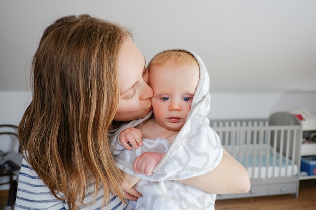 Jovem mãe segurando e beijando o bebê seco, usando uma toalha com capuz, após o banho ou duche. Vista frontal. Conceito de cuidado infantil ou banho