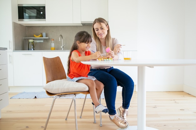 Jovem mãe segurando ovo de páscoa, sorrindo e mostrando a filha na cozinha.