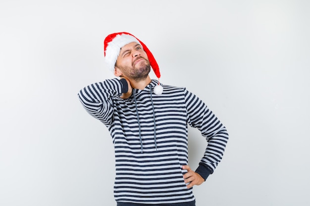 Jovem mantendo a mão no pescoço, olhando para cima com um capuz, chapéu de papai noel e parecendo pensativo,