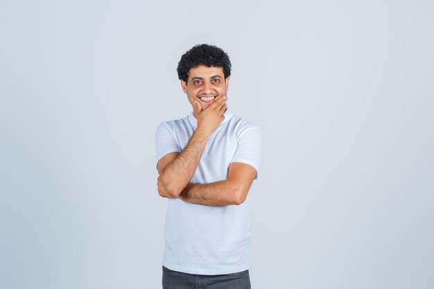 Jovem, mantendo a mão no queixo em camiseta branca, calça e parecendo alegre. vista frontal.