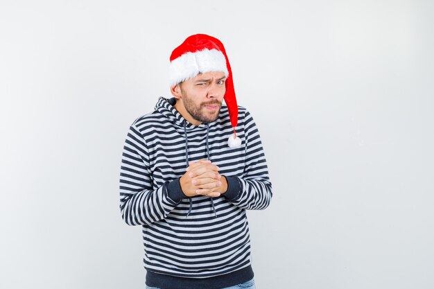 Foto grátis jovem, mantendo as mãos cruzadas sobre o peito com capuz, chapéu de papai noel e parecendo curioso.