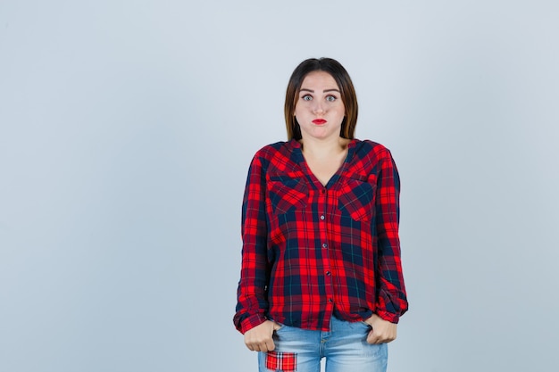 Jovem, mantendo os polegares nos bolsos, enquanto assoando as bochechas em uma camisa xadrez, jeans e parecendo entediada, vista frontal.