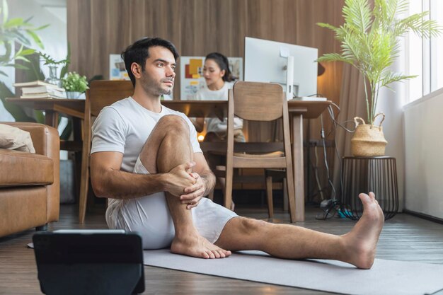 Jovem marido asiático treina exercícios de ioga praticando aula on-line feomr na sala de estar com a esposa trabalhando atrás do conceito de ordem de estado de casa ficar em casa