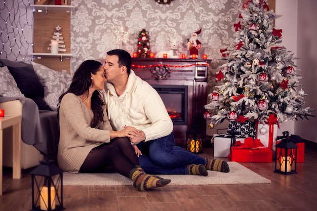 Jovem marido beijando a testa da esposa no dia de Natal em frente à lareira acolhedora. Comemorando o natal.