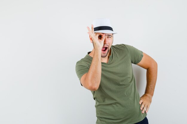 Jovem masculino com camiseta e chapéu verdes, jeans mostrando sinal de ok no olho e parecendo engraçado