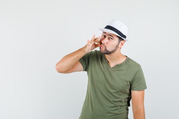 Jovem masculino com camiseta verde e chapéu mostrando gesto de zíper e parecendo seguro