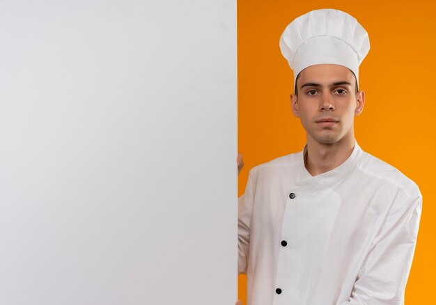 Foto grátis jovem masculino legal vestindo uniforme de chef segurando uma parede branca na parede amarela isolada com espaço de cópia