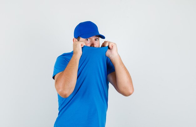 Jovem masculino puxando a gola do rosto com camiseta azul e boné e parecendo assustado