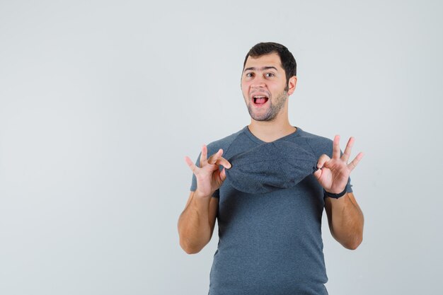 Jovem masculino segurando um boné em uma camiseta cinza e parecendo brincalhão