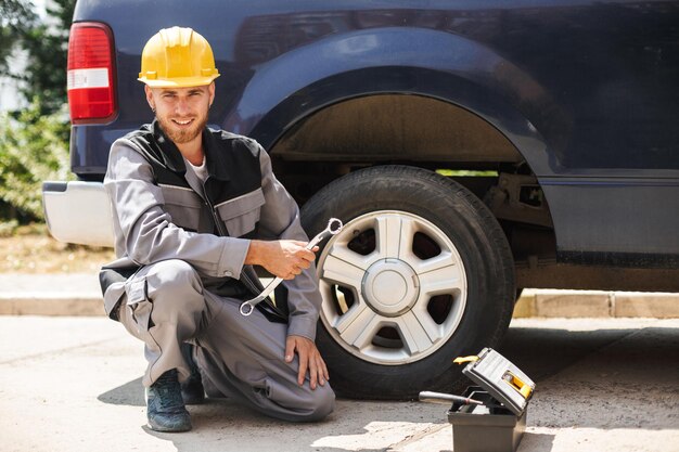 Jovem mecânico sorridente em roupas de trabalho e capacete amarelo olhando alegremente na câmera segurando a chave para trocar a roda do carro ao ar livre