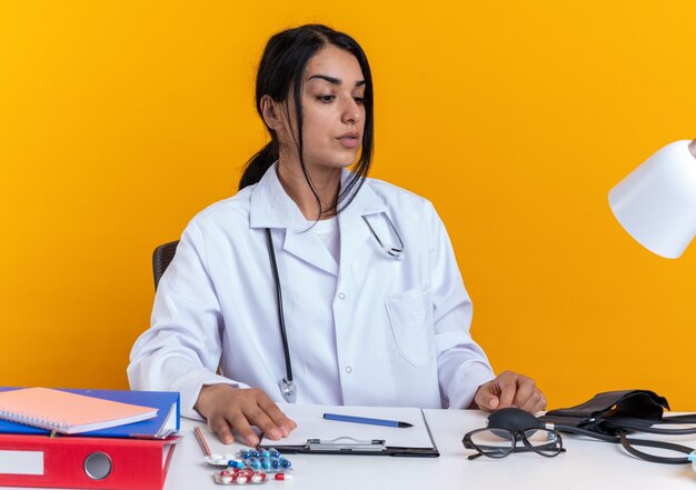 Jovem médica a pensar, vestindo manto médico com estetoscópio, se senta à mesa com ferramentas médicas isoladas em fundo amarelo