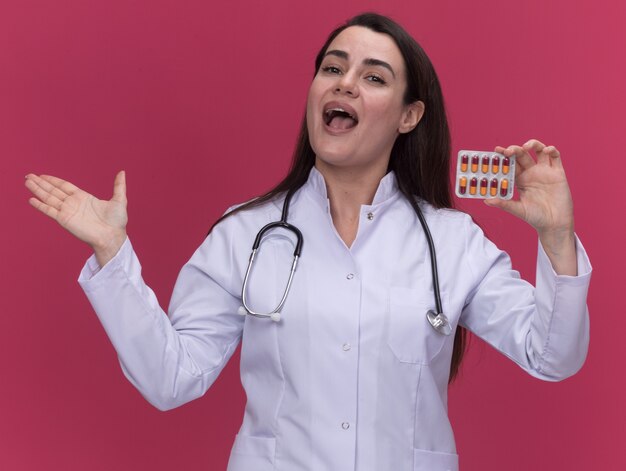 Foto grátis jovem médica alegre vestindo bata médica com estetoscópio e mantendo a mão aberta