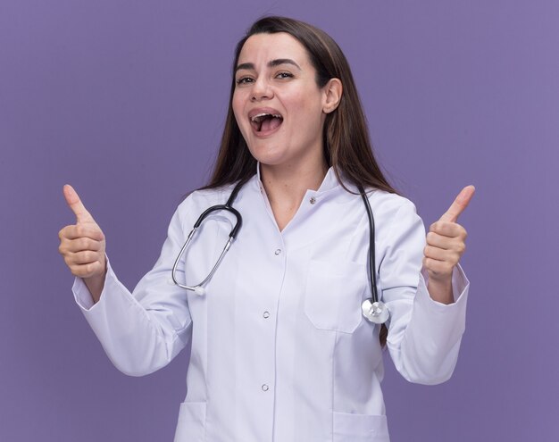 Jovem médica alegre vestindo túnica médica com estetoscópio polegar para cima e duas mãos isoladas na parede roxa com espaço de cópia