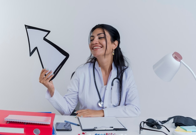 Jovem médica alegre vestindo túnica médica e estetoscópio, sentada à mesa com ferramentas médicas, segurando e olhando para a marca de seta apontando para o lado isolado