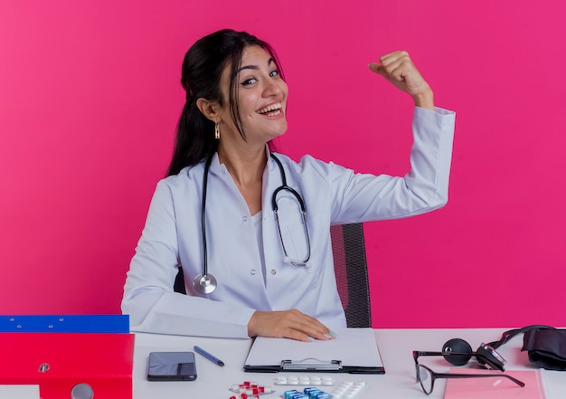 Jovem médica alegre vestindo túnica médica e estetoscópio sentada na mesa com ferramentas médicas, colocando a mão na mesa fazendo um gesto forte isolado na parede rosa