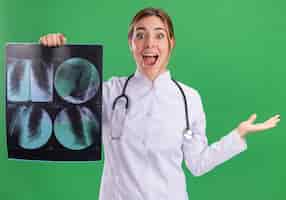 Foto grátis jovem médica animada vestindo túnica de médico com estetoscópio segurando um raio-x estendendo a mão isolada na parede verde