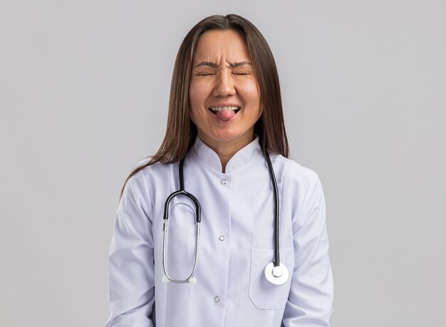 Jovem médica asiática, brincalhona, vestindo bata médica e estetoscópio, mostrando a língua com os olhos fechados, isolada na parede branca