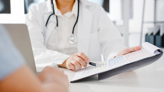 Jovem médica asiática em uniforme médico branco, usando a prancheta, está dando ótimas notícias.