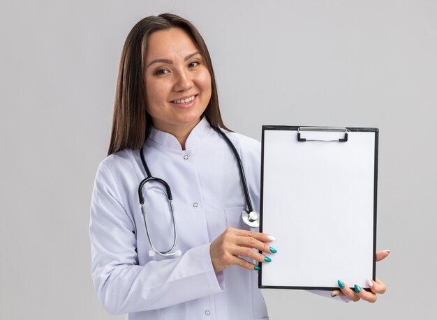 Jovem médica asiática sorridente usando túnica médica e estetoscópio mostrando a área de transferência para a câmera, olhando para a frente, isolada na parede branca