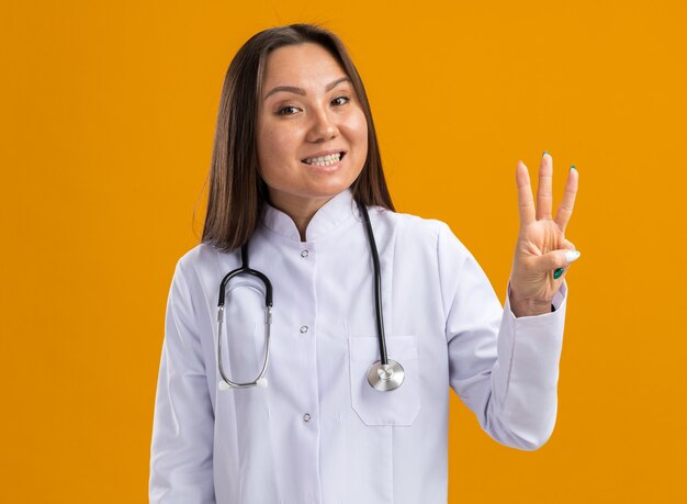 Jovem médica asiática vestindo bata médica e estetoscópio, olhando para a frente, mostrando os dentes e três com a mão isolada na parede laranja