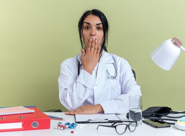 Jovem médica assustada vestindo túnica médica com estetoscópio sentada na mesa com ferramentas médicas cobrindo a boca com a mão isolada no fundo verde oliva