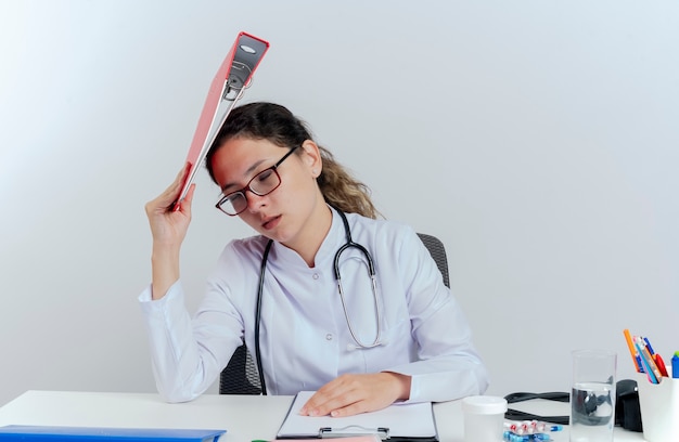 Jovem médica cansada vestindo túnica médica, estetoscópio e óculos, sentada na mesa com ferramentas médicas, tocando a cabeça com a pasta com os olhos fechados isolados