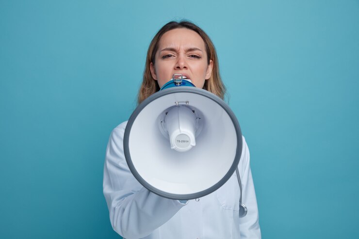 Foto grátis jovem médica carrancuda usando roupão médico e estetoscópio pendurado no pescoço falando pelo alto-falante