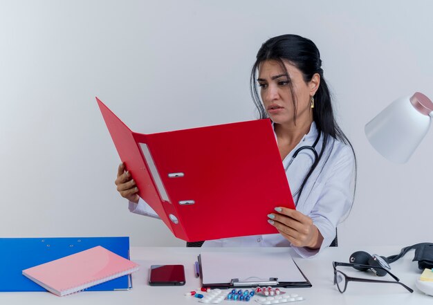 Jovem médica carrancuda, vestindo túnica médica e estetoscópio, sentada à mesa com ferramentas médicas, segurando e olhando para a pasta isolada