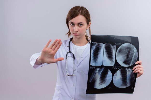 Jovem médica com aparência séria, vestindo bata médica e estetoscópio, segurando uma foto de raio-x, fazendo um sinal de pare